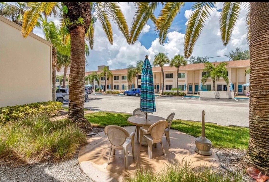 A shaded outdoor seating area is created under a large umbrella, featuring a round table surrounded by four chairs. Lush greenery and palm trees frame the space, while a pathway leads to the low-rise building in the background.