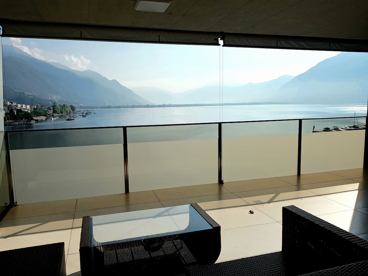 A spacious balcony is presented with a glass railing, offering unobstructed views of the lake and surrounding mountains. Modern outdoor furniture, including a coffee table, is positioned on the tiled floor, creating an inviting space for relaxation. Soft natural light bathes the area.
