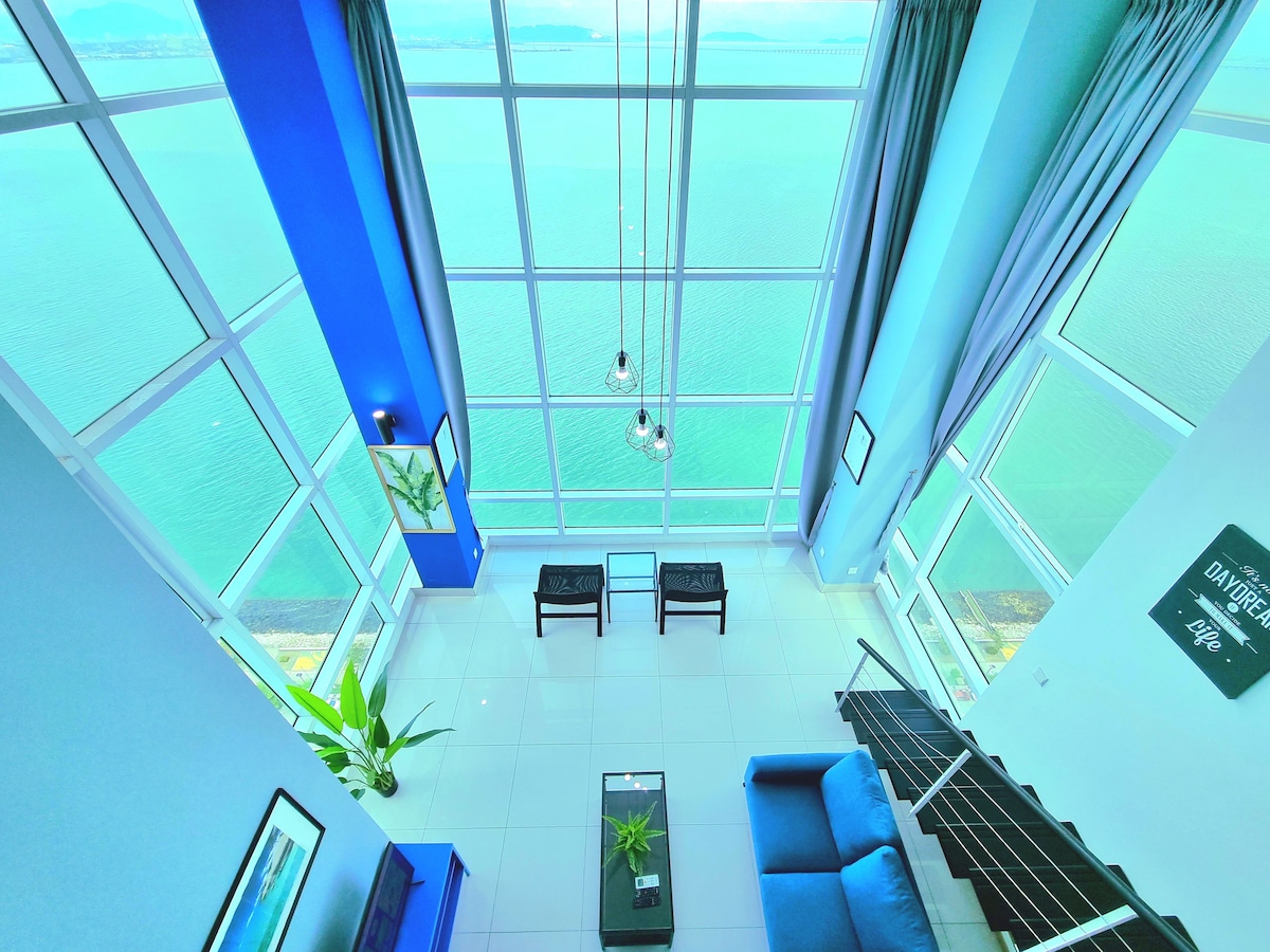 A bright, open living space features floor-to-ceiling windows, offering expansive views of the sea. Two chairs are positioned next to a glass coffee table, with a blue sofa inviting relaxation. The walls are painted in light blue and white, enhancing the airy feel of the room.