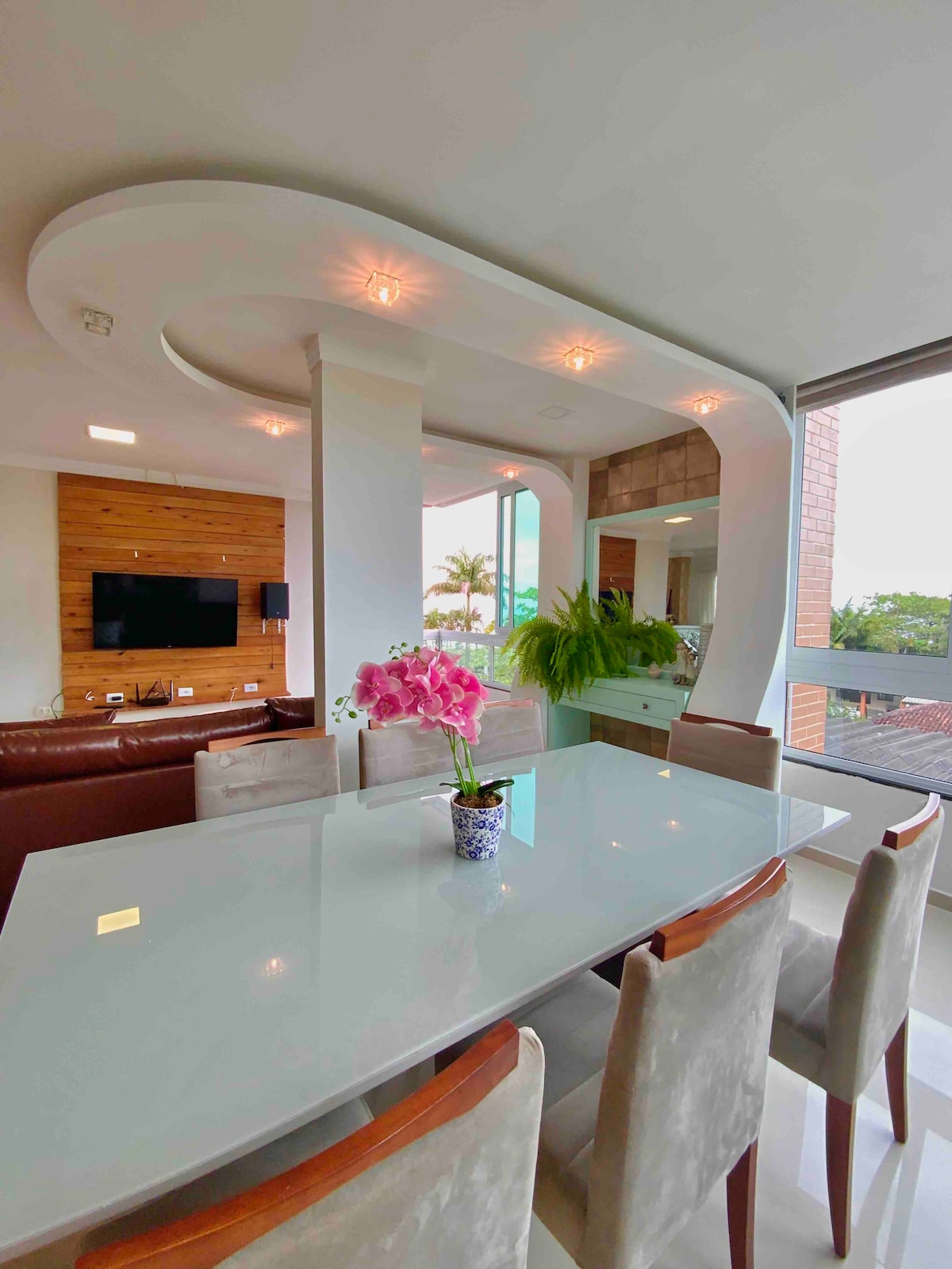 A light-filled dining area features a sleek white table surrounded by upholstered chairs. A decorative vase holding purple orchids sits in the center. A modern living space with a television is visible in the background, framed by large windows offering views of the outdoors.