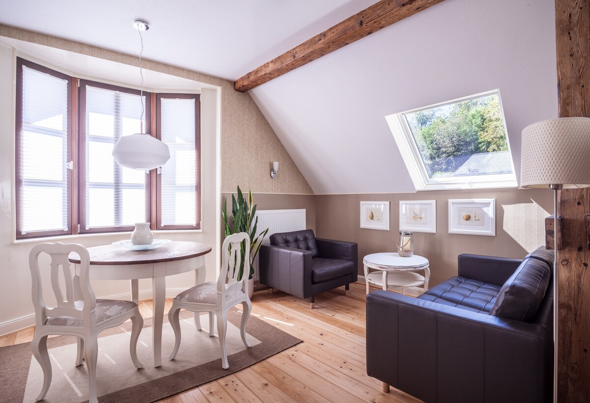 A bright living area features a round dining table with four chairs beside a cozy seating arrangement consisting of two black armchairs and a small white coffee table. Natural light streams through large windows, while wooden beams add character to the sloped ceiling.