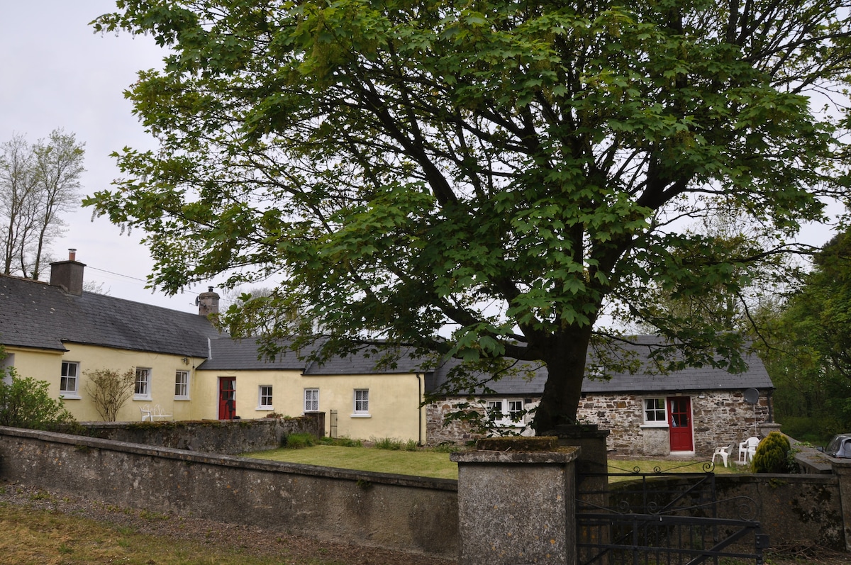 A charming cottage exterior is shown, featuring yellow walls and red doors. A large tree provides shade over the private garden area, which is bordered by a low stone wall. The surrounding greenery enhances the tranquil ambiance of the property.