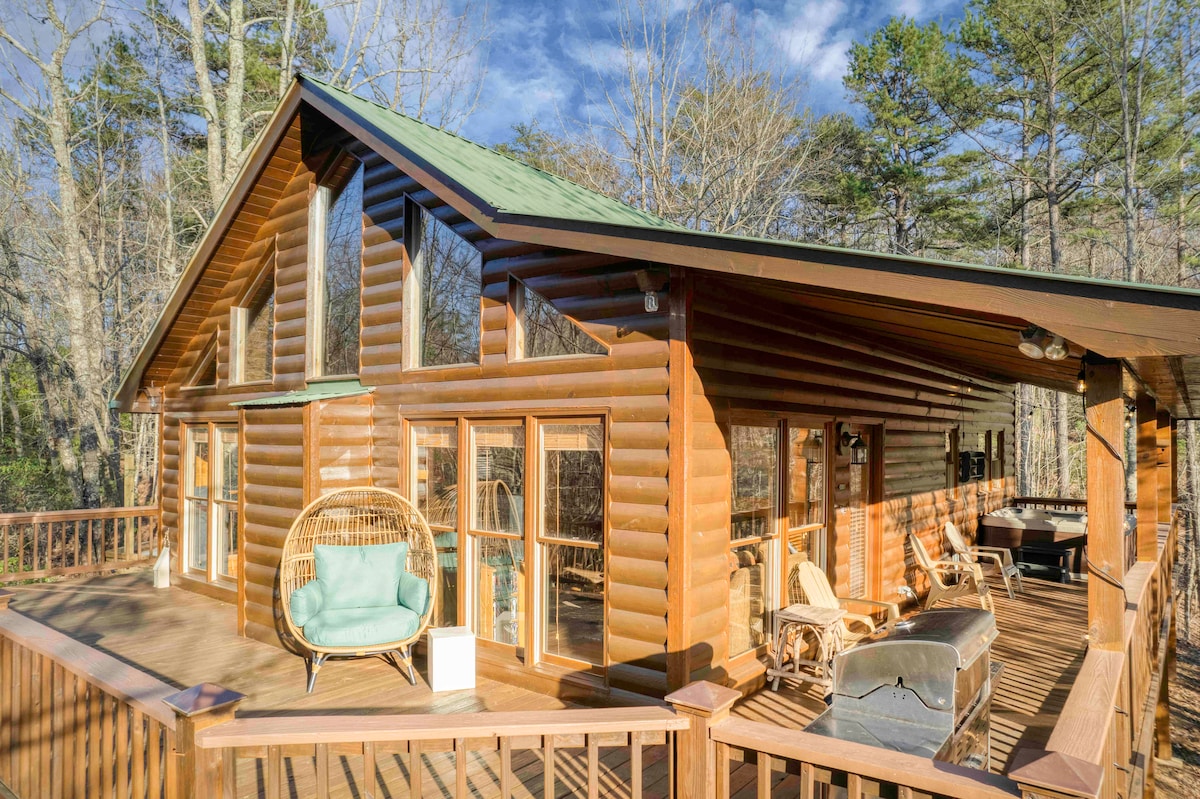 A log cabin exterior is showcased, featuring large windows and a unique, modern design. A spacious deck surrounds the cabin, furnished with a swing chair and relaxing seating options. The natural surroundings complement the tranquil setting, inviting guests to enjoy the outdoor space.