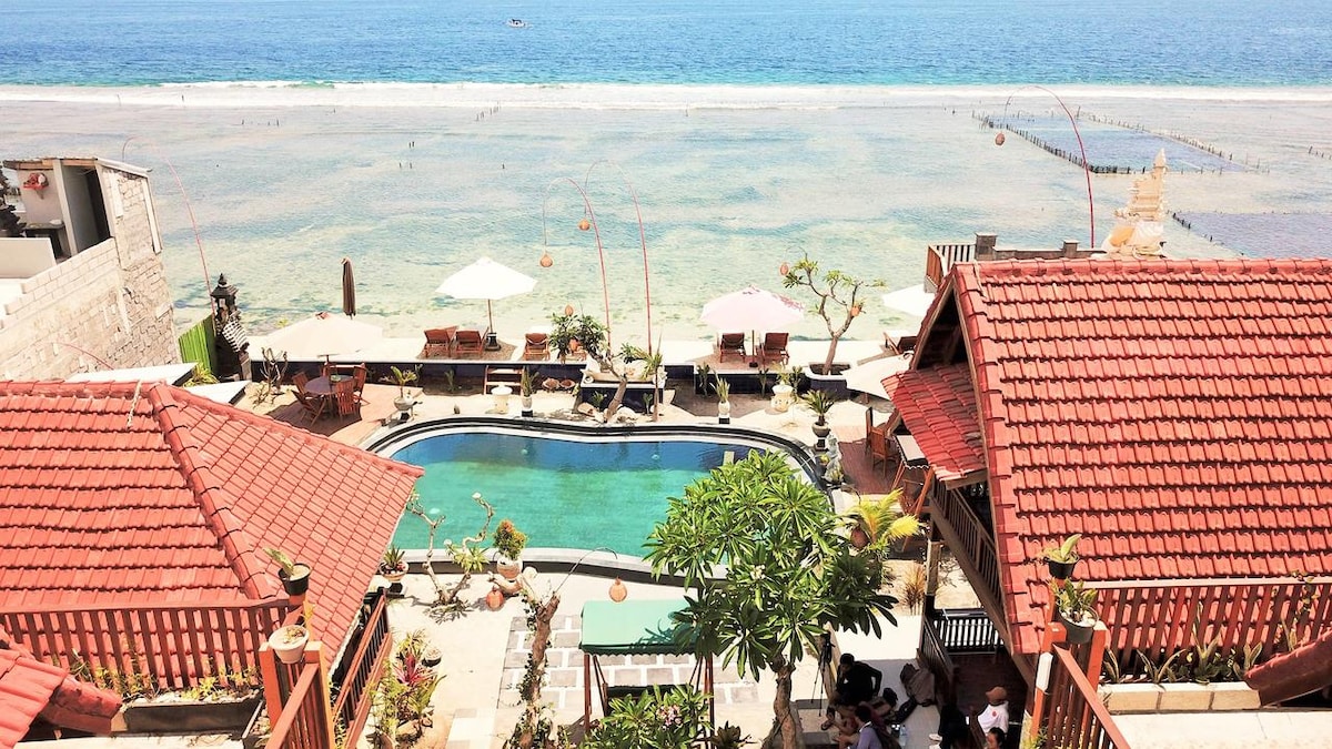 The image displays a resort featuring a clear swimming pool surrounded by lush greenery and lounge chairs. Traditional structures with red-tiled roofs line the area, overlooking a serene beach with calm water. Umbrellas provide shade for relaxation, enhancing the inviting outdoor atmosphere.