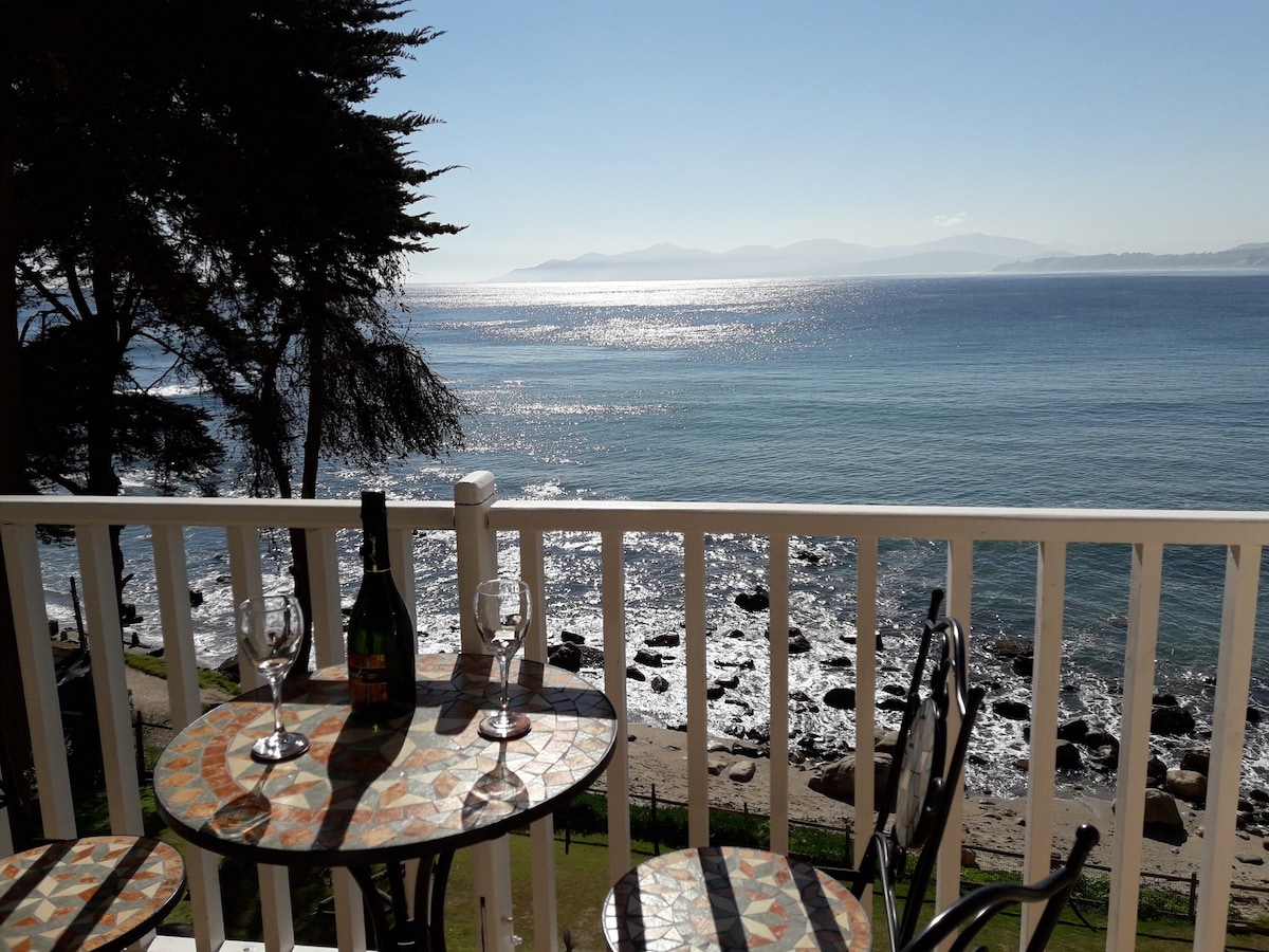A balcony table adorned with a mosaic surface holds a bottle and two glasses, overlooking gently rippling water. The serene view includes the coastline and distant mountains under a clear blue sky, enhancing the tranquil setting.