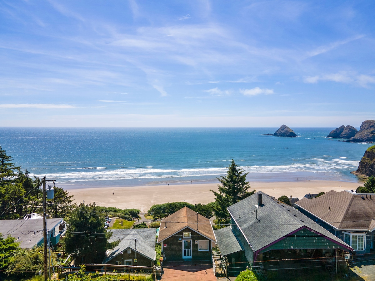 A panoramic view of the sandy beach and ocean, with gentle waves lapping at the shore. The distant Three Arch Rocks are visible against the shimmering water, framed by lush greenery and residential structures. The sky is bright and clear, enhancing the serene coastal atmosphere.