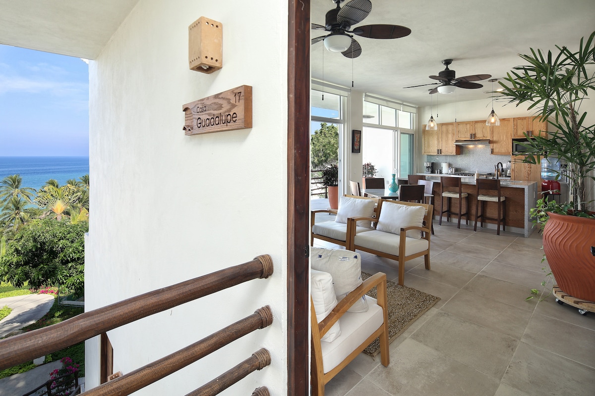 An entrance view displays a bright and airy living space with a cozy seating area and kitchen. Lush greenery is featured in the background, while expansive windows allow for natural light and a stunning ocean view. The sign indicates the property name, Condo Guadalupe.
