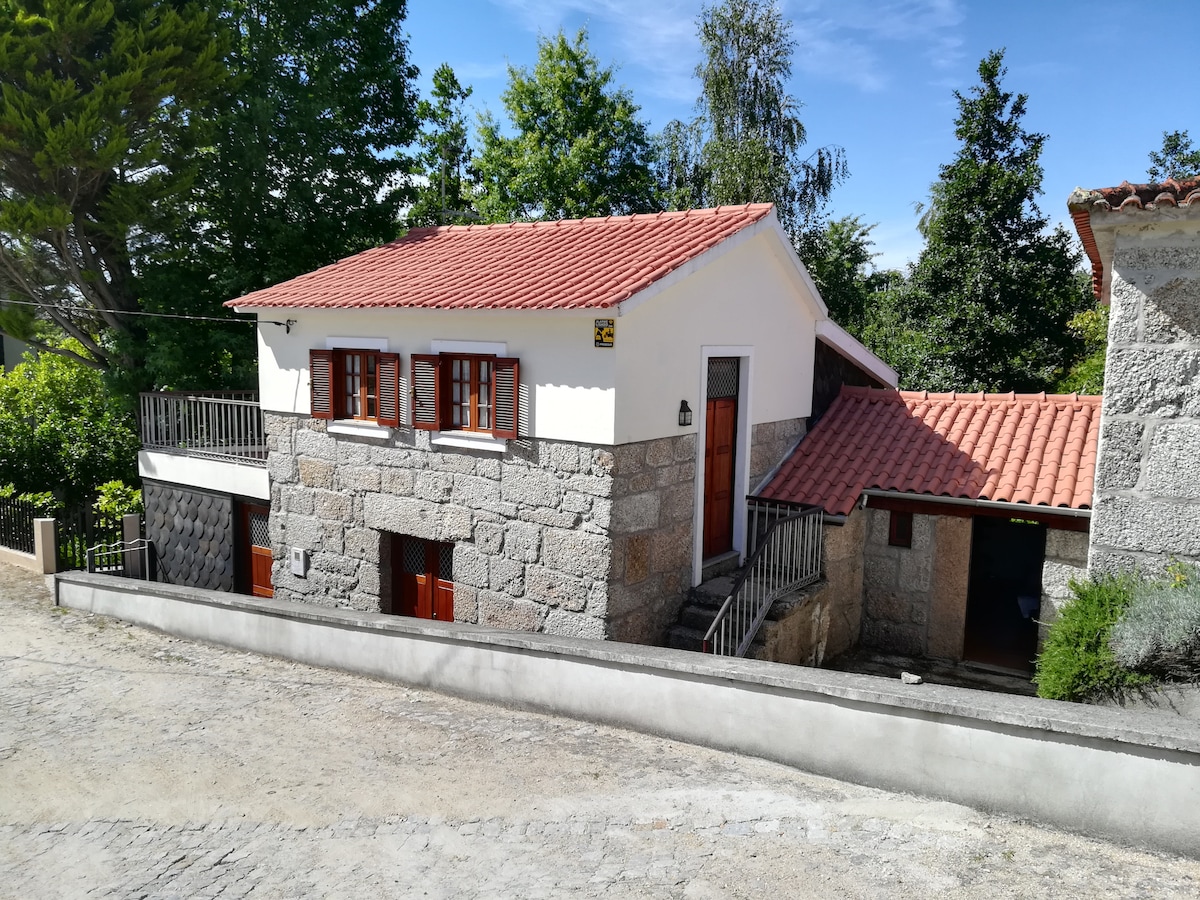 A rustic stone house features a sloped roof with red tiles and wooden windows. The structure has a warm exterior, complemented by greenery surrounding it. A simple pathway leads up to the entrance, offering easy access to the inviting atmosphere of the home.