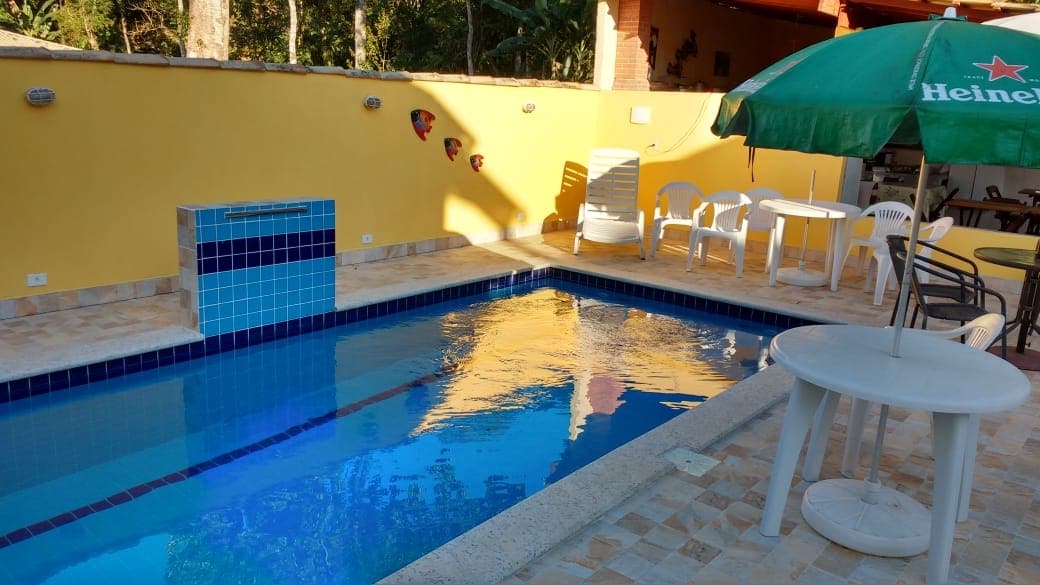 A refreshing swimming pool occupies the outdoor space, featuring a blue-tiled accent wall. Surrounding the pool are several white chairs and tables, along with a large green umbrella for shade. A bright yellow wall creates a vibrant backdrop, enhancing the inviting setting.