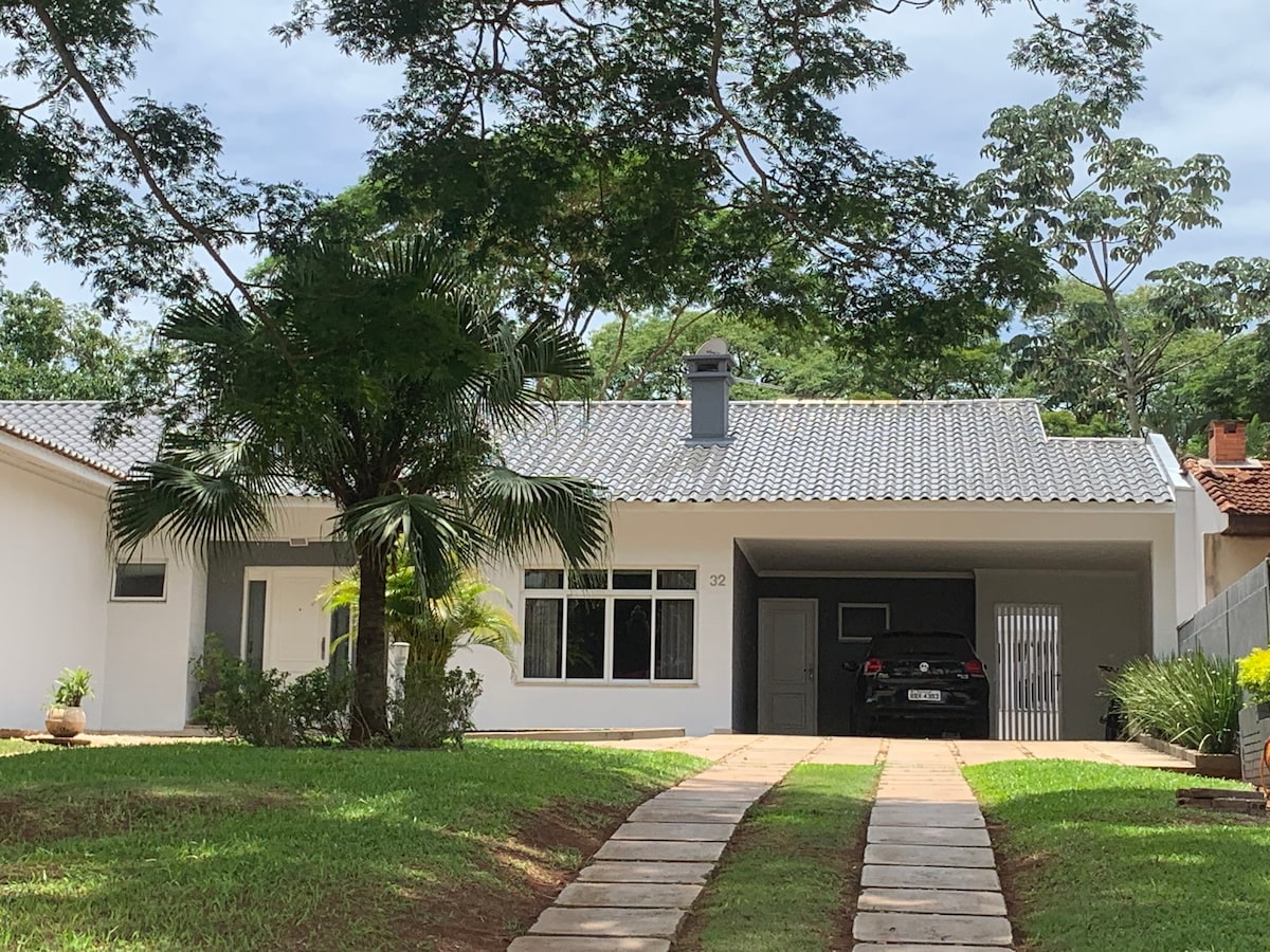 The exterior of a modern single-story house is depicted, featuring a grey tiled roof and a large front yard. A driveway leads to a covered parking space, while strategically placed palm trees and vibrant greenery enhance the tranquil atmosphere.