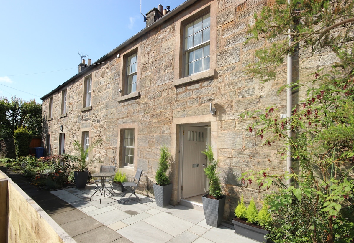 The exterior of the cottage features stone walls and a private entrance. A small patio area is furnished with a bistro table and chairs, surrounded by potted plants. The garden is enclosed and offers a peaceful escape.