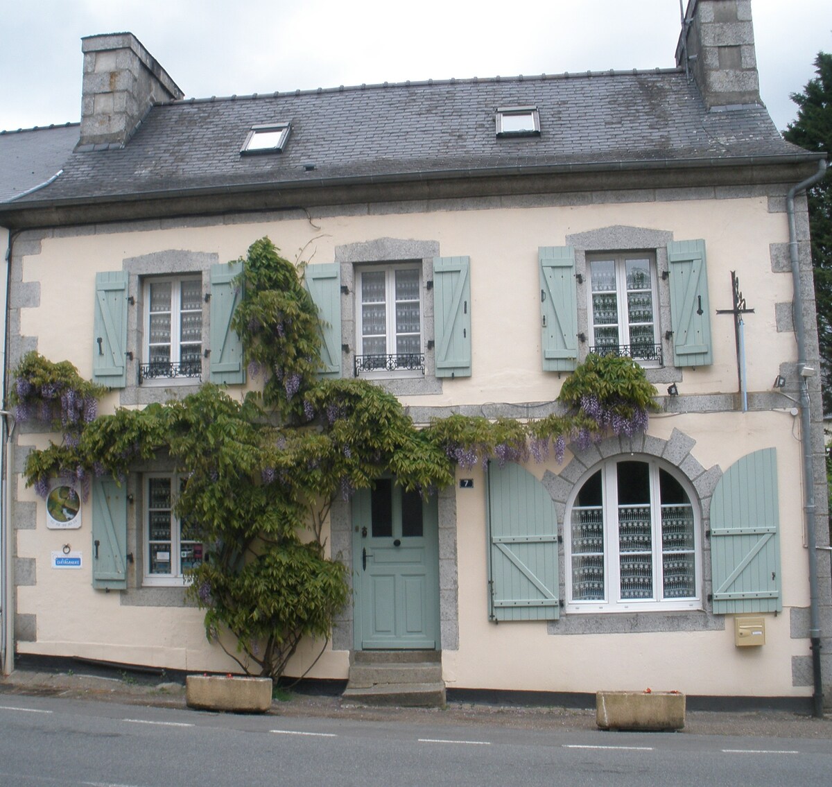 A charming building features a light-colored exterior with green shutters and a central door. Wisteria vines gracefully climb over the facade, adding a natural touch. The windows are adorned with decorative grills, providing a glimpse of the character inside.
