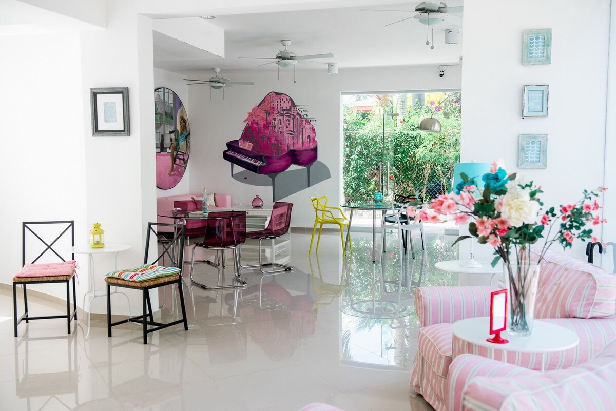 A bright and airy common area features modern furniture arrangements, including colorful chairs and a sleek dining table. A large mural depicts a vibrant scene, adding a pop of color, while large windows invite natural light and views of greenery from outside.