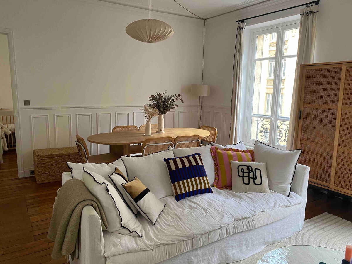 A bright living and dining area features a white sofa adorned with textured cushions. A round wooden dining table is surrounded by several chairs. Natural light filters through large windows, illuminating the space. A stylish lamp and woven storage unit are visible in the background.