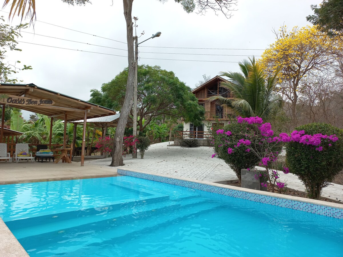 A refreshing pool is seen in the foreground, bordered by lush tropical trees and vibrant flowering plants. In the background, a beautifully constructed chalet is visible, complemented by a shaded outdoor seating area under a roof, creating a serene atmosphere.
