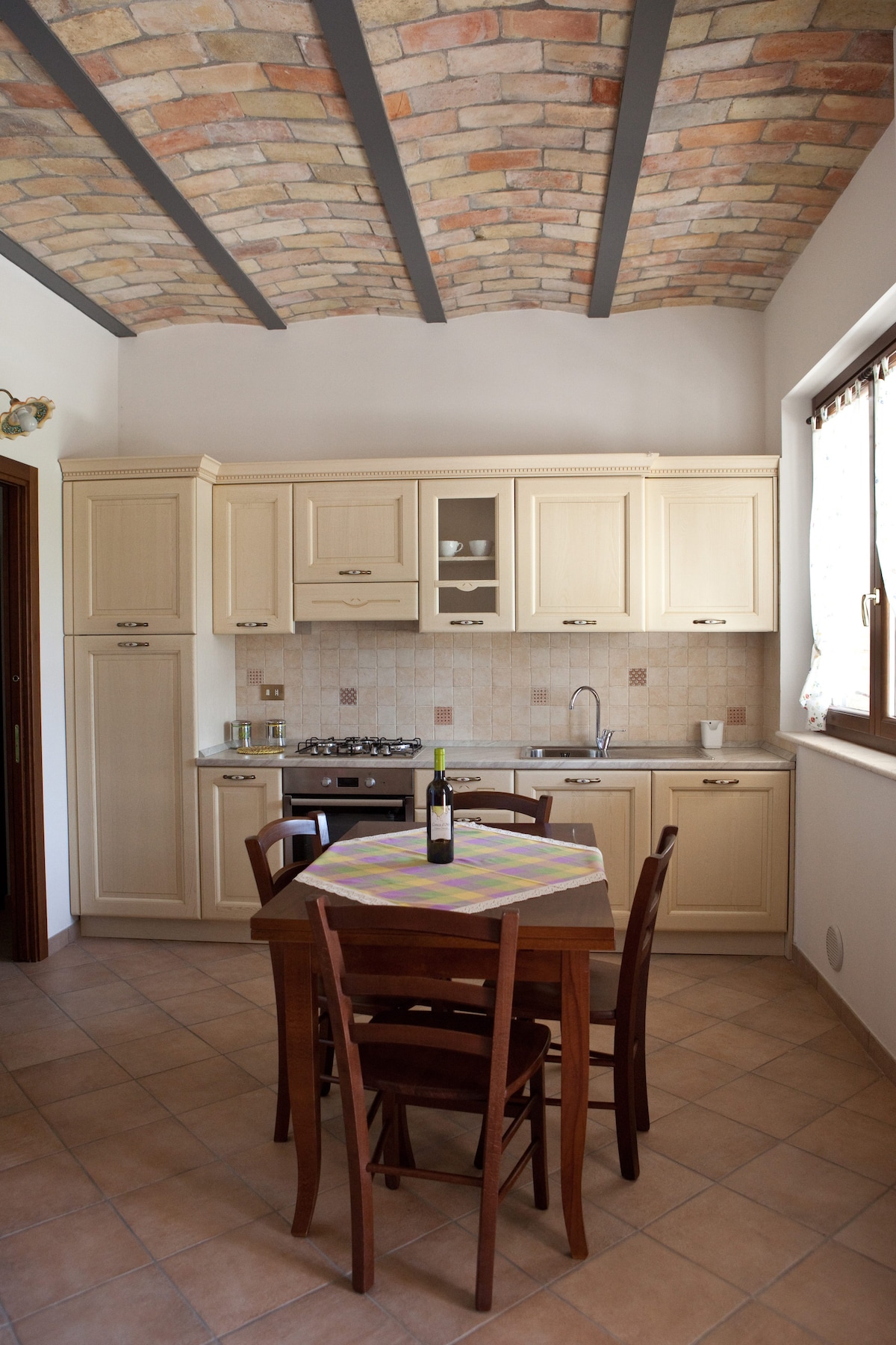 The kitchen features light-colored cabinetry and a gas stove, complemented by a neutral backsplash. A wooden dining table with four chairs is centered in the space. A bottle of wine sits on the table, while natural light filters through a window adorned with patterned curtains.