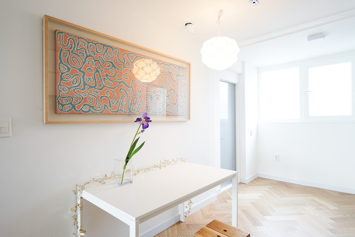 The dining area features a white table adorned with a simple glass vase holding a purple flower. A contemporary artwork, framed in light wood, hangs above the table. Light fixtures provide illumination, while large windows bring natural light into the space.