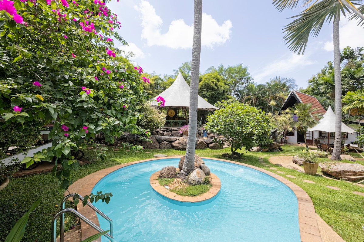 A tranquil outdoor setting is showcased with a curved swimming pool surrounded by lush greenery. Tropical plants, including vibrant flowering bushes, frame the scene. A large palm tree rises above, enhancing the natural ambiance of the well-kept garden area.