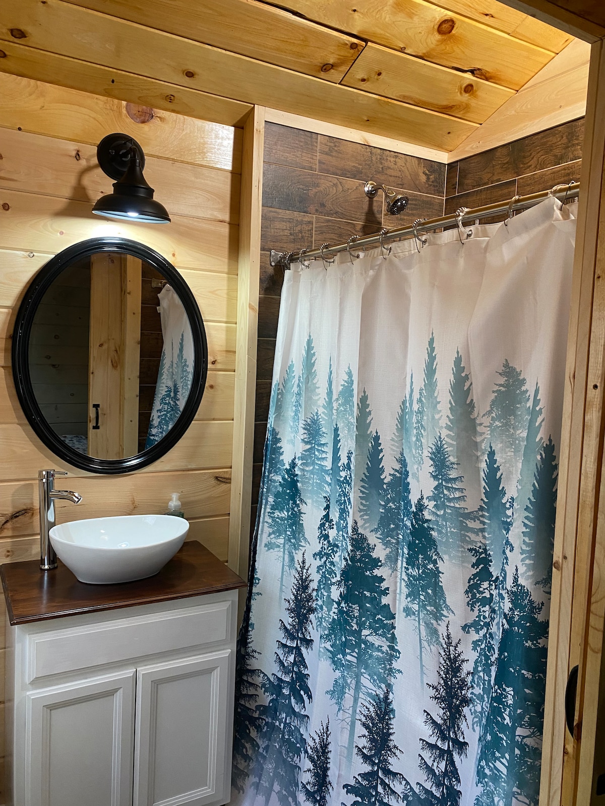 A serene bathroom features warm wooden walls and a modern sink on a wooden vanity. A round mirror is mounted above the sink, and a unique shower curtain displays a forest scene with tall evergreen trees, enhancing the cabin's natural ambiance.