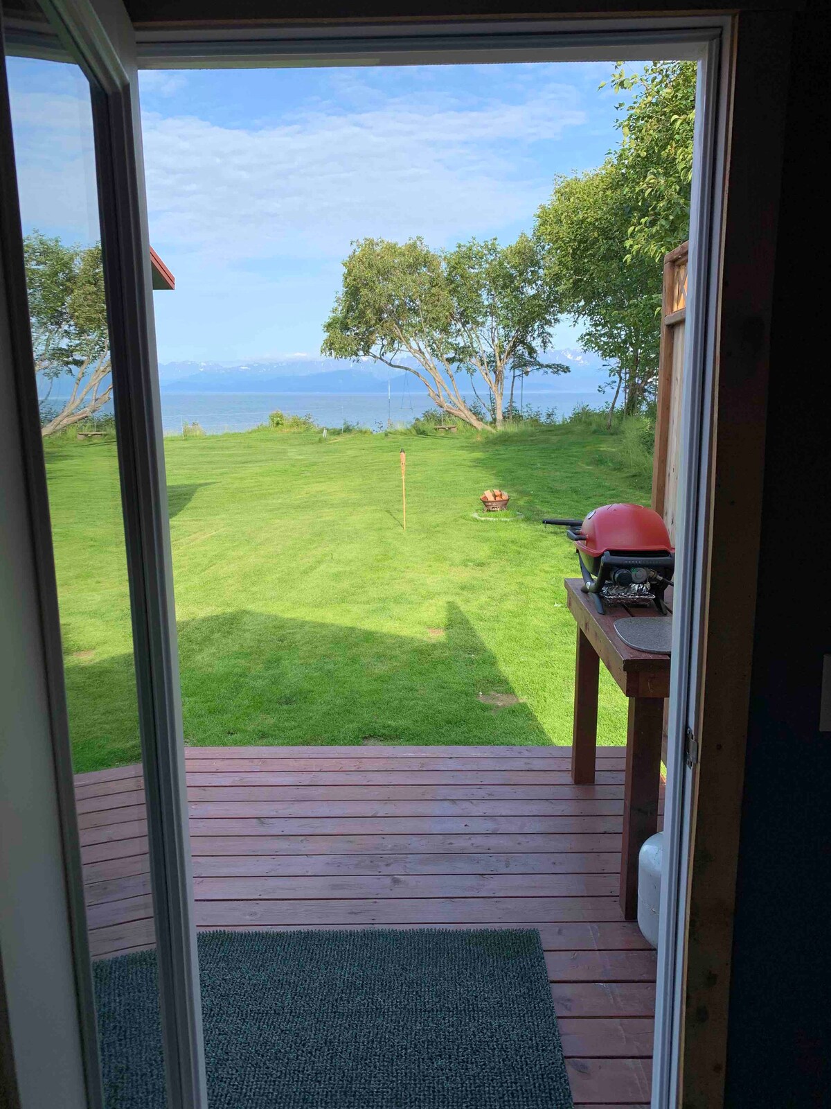 A view from an open sliding door reveals a well-maintained lawn leading to a serene waterfront. A small grill is positioned on a wooden deck, providing an inviting outdoor space. Lush greenery and a clear sky enhance the peaceful ambiance.
