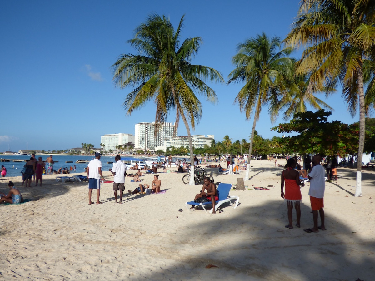 A sandy beach scene features palm trees swaying gently in the breeze. Groups of people enjoy the sun, some resting on lounge chairs, while others walk along the shore. In the background, resort buildings provide a glimpse of nearby accommodations.