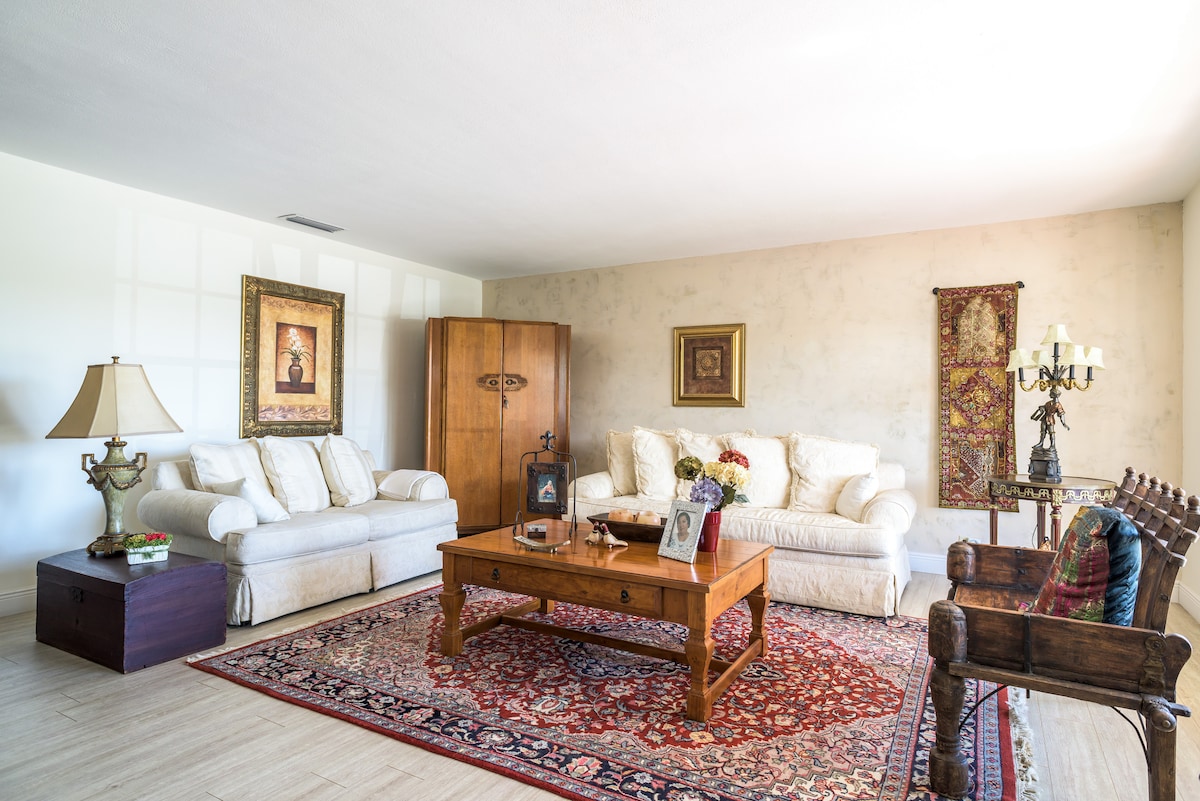 A spacious living area features two light-colored sofas and a wooden coffee table set on a patterned area rug. Decorative artwork is displayed on the walls. A vintage wooden cabinet and a candleholder table lamp add character to the serene environment.