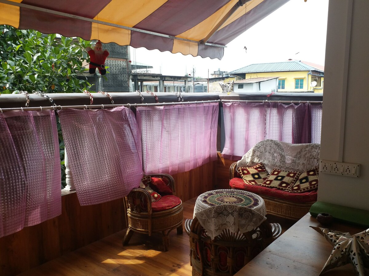 A cozy balcony area features woven wicker chairs adorned with colorful cushions. A round table is positioned in the center, complemented by a lace tablecloth. Sheer purple curtains frame the space, creating a relaxed atmosphere, while greenery is visible in the background.