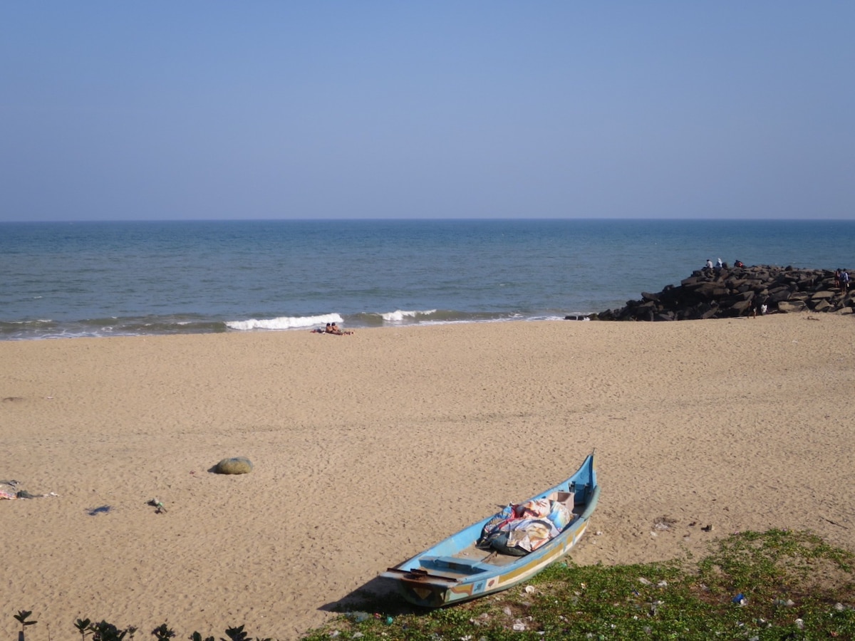 A sandy shoreline stretches along the Bay of Bengal, with gentle waves lapping at the beach. A blue wooden fishing boat rests in the foreground, while a rocky outcrop can be seen in the distance. The clear sky enhances the serene coastal view.