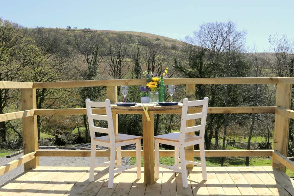 A wooden deck features a small round table set for two with a vase of flowers, overlooking scenic hills and tree-lined views. Two white chairs provide comfortable seating, enhanced by natural sunlight illuminating the space.