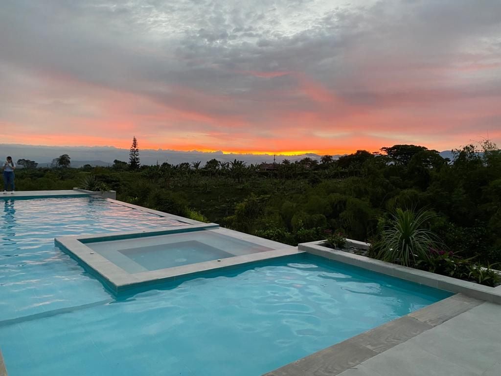 An infinity pool stretches into the landscape, reflecting shades of blue. A jacuzzi is seen nestled within the pool's edge. In the background, a vibrant sunset displays hues of orange and pink against a softly clouded sky, framed by lush greenery.