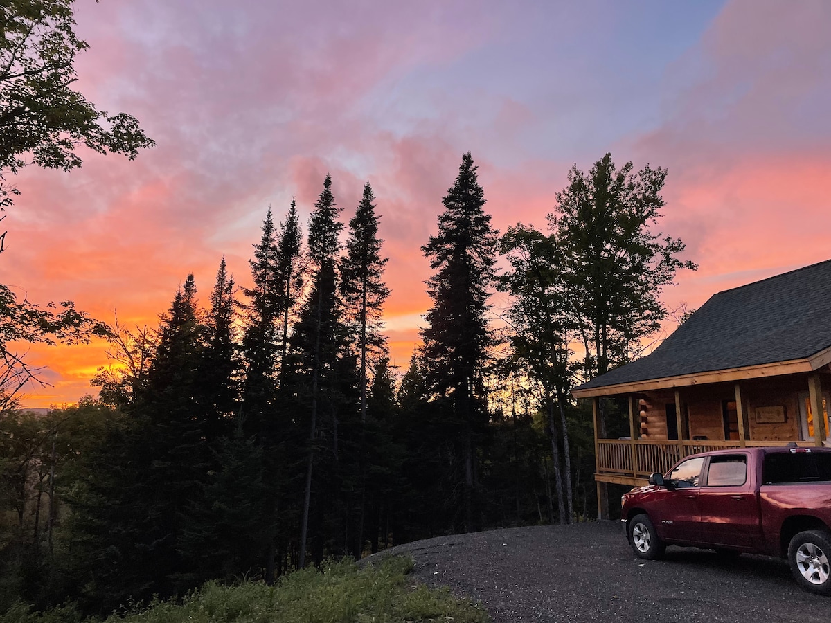 A tranquil evening scene showcases a log cabin, partially obscured by tall evergreen trees. A vehicle is parked on a gravel area in the foreground. The sky is painted with vibrant pink and orange hues, creating a serene backdrop to the setting.
