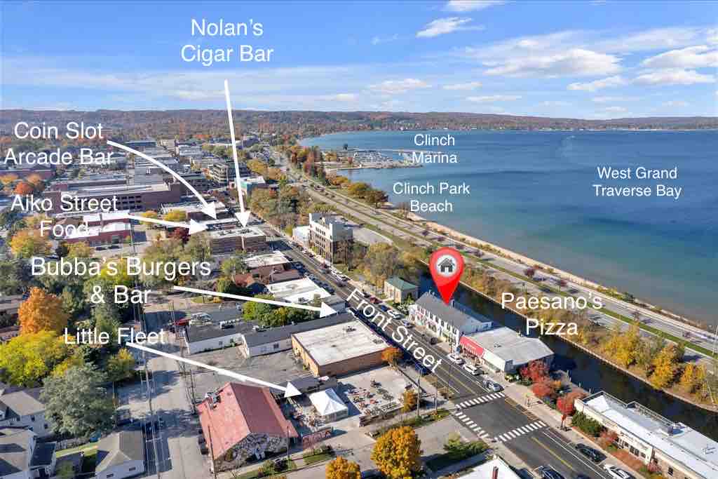 An aerial view captures the vibrant downtown Traverse City against a stunning backdrop of West Grand Traverse Bay. Key local establishments are highlighted, showcasing the area’s accessibility to dining and recreation. The marina and beach visible in the distance contribute to the inviting atmosphere of the location.