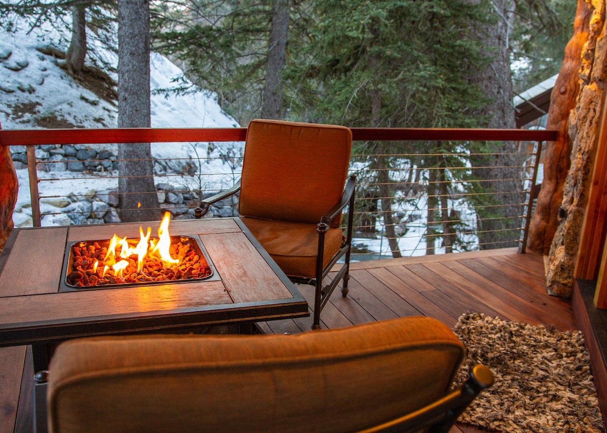 A cozy outdoor seating area features two armchairs positioned around a fire pit. The warm flames provide a contrast to the snowy backdrop of trees and a flowing stream, creating a serene atmosphere for relaxation.