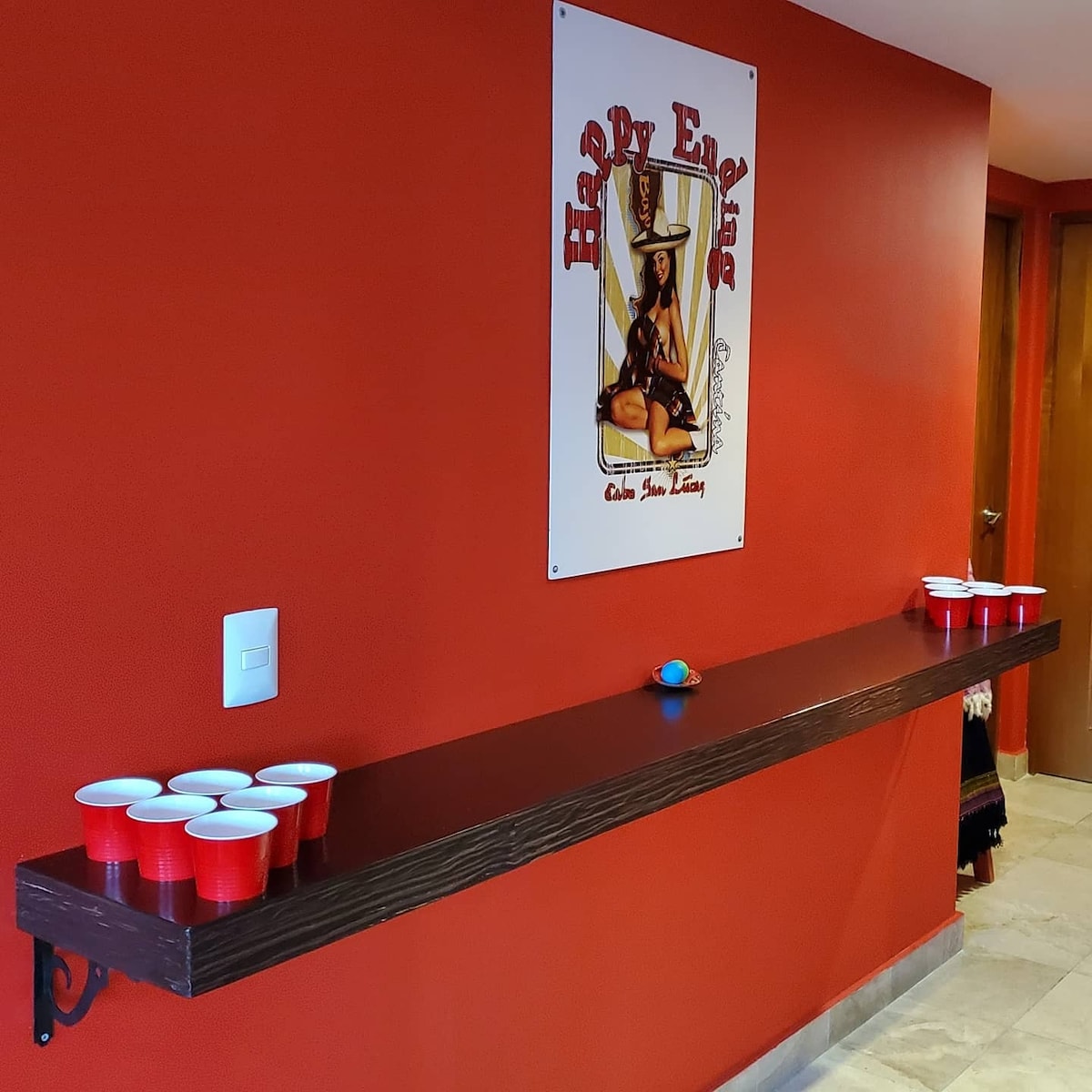 A vibrant red wall is displayed with a piece of artwork framed above a wooden ledge. The ledge features a row of red cups arranged neatly, suggesting a casual games area. The light-colored flooring contrasts softly with the bold wall color.