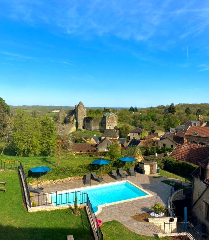 A view of a serene outdoor space featuring a heated swimming pool surrounded by lounge chairs and umbrellas. The backdrop includes historical stone ruins and lush greenery, enhancing the setting's natural beauty under a clear blue sky.