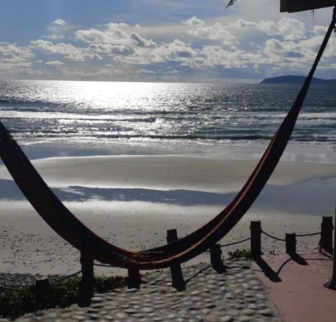 A hammock hangs serenely near the water's edge, overlooking a wide expanse of shimmering ocean. Soft waves lap against the sandy shore, while clouds drift across a bright sky, creating a tranquil seaside setting.
