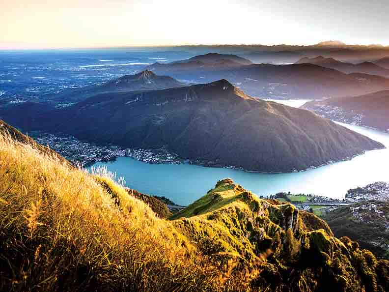 A panoramic view captures the mountainous landscape surrounding Lake Como. Rolling hills covered in green grass meet the water's edge, with sunlight illuminating the peaks and valleys. The distance showcases towns nestled along the lake, contributing to a serene natural setting.