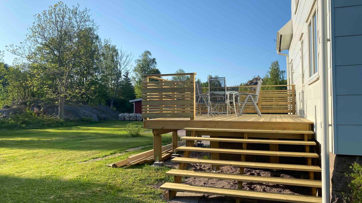 A wooden deck extends from the house, featuring a set of white outdoor chairs and a table. A grassy area is visible nearby, surrounded by trees and rocks. The landscape is bathed in natural light, presenting a serene outdoor space for relaxation.