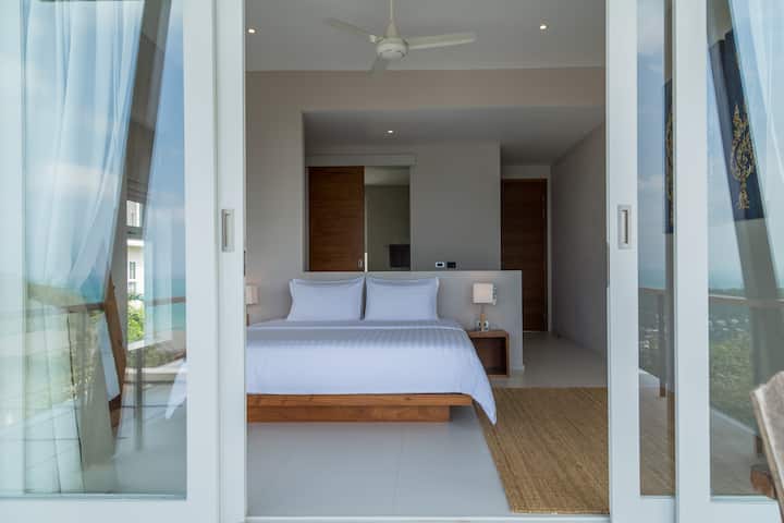 Елегантна спалня с огромно двойно легло и плъзгащи се врати, които се отварят към морски гледки, естествена светлина и изчистен модерен дизайн.