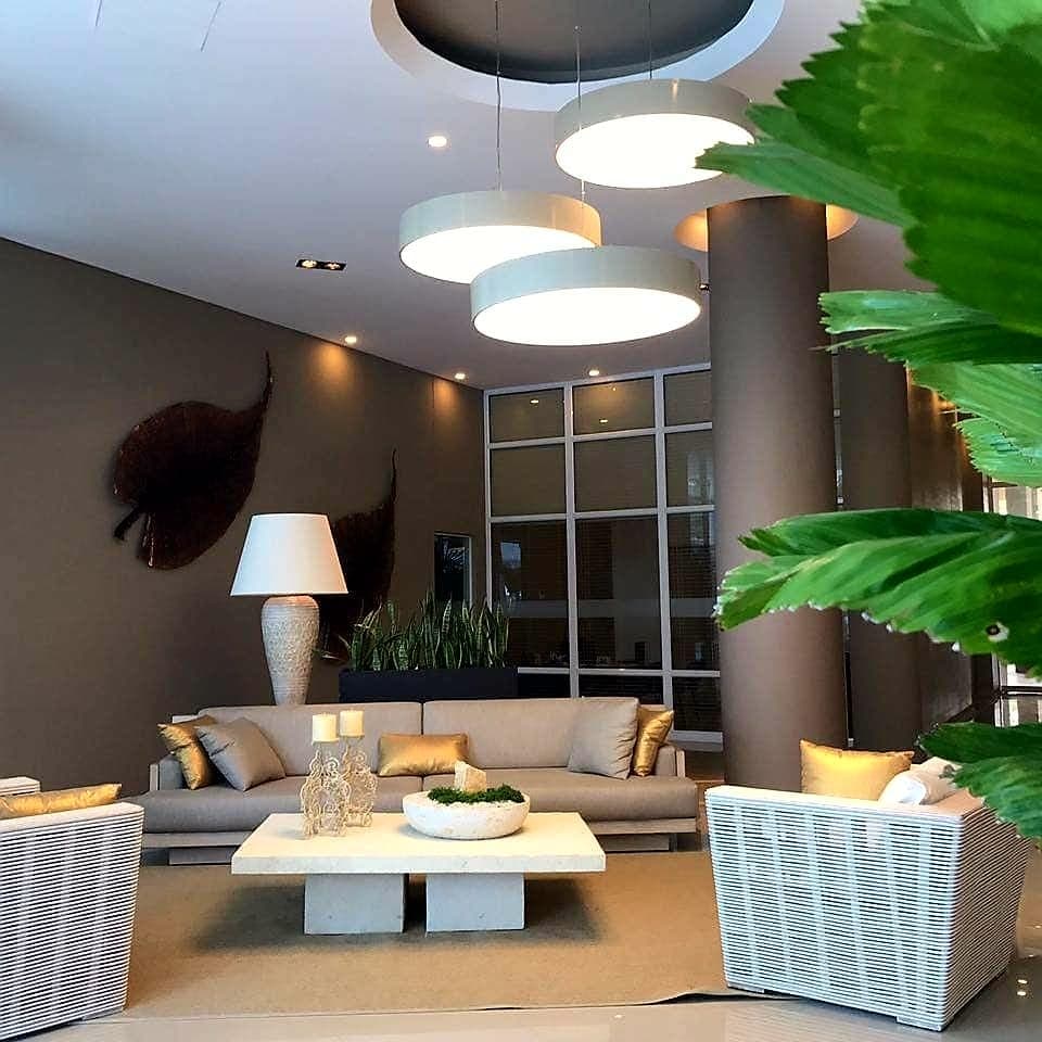 A modern lobby is showcased, featuring a plush sofa and two woven chairs surrounding a coffee table. Soft lighting is provided by three pendant lights above, while two decorative plants enhance the inviting atmosphere.