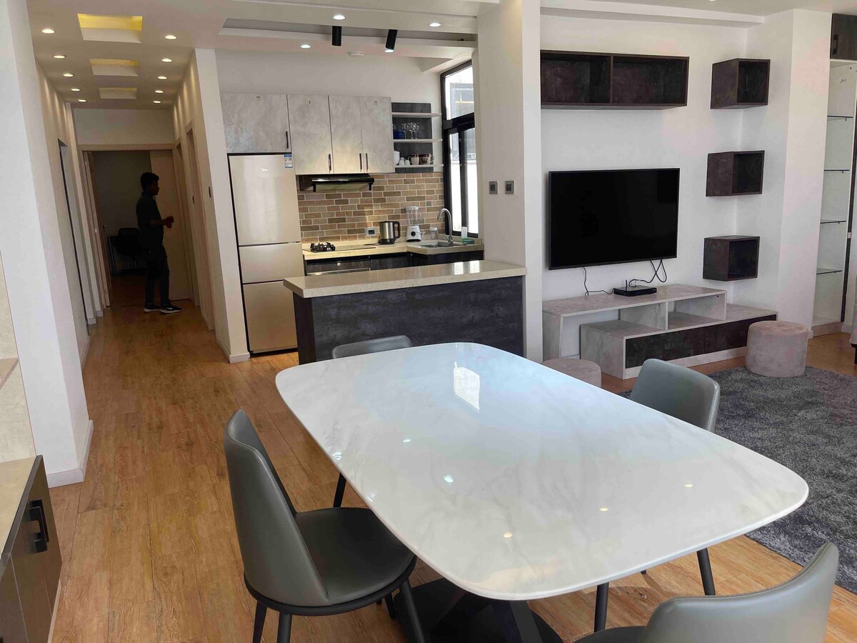 A spacious living area features a modern dining table surrounded by several chairs. An open kitchenette is visible with sleek cabinetry and appliance arrangements. A wall-mounted television and shelving units are positioned in the background. Natural light enters through large windows.