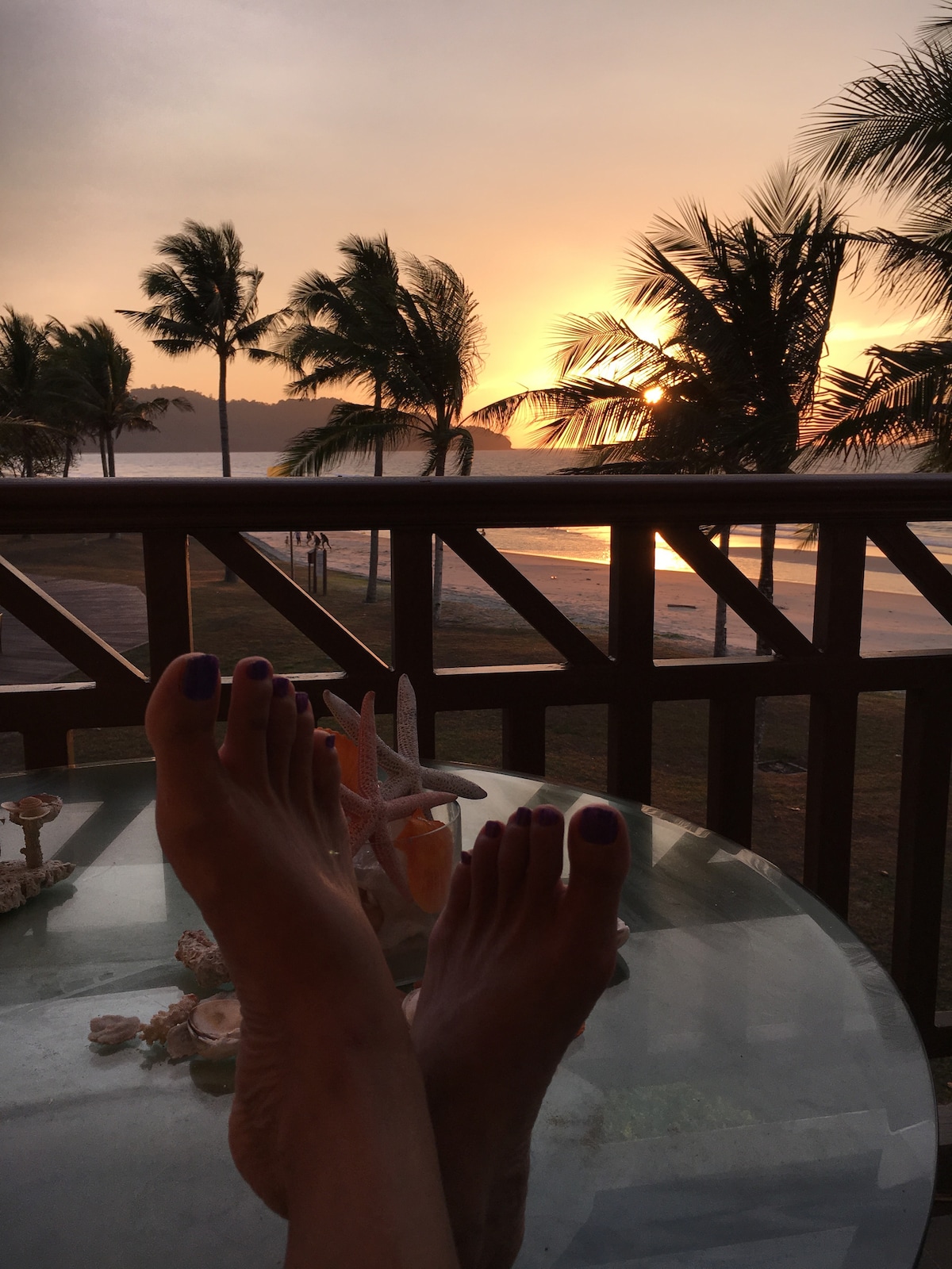 A balcony view captures a serene sunset, with palm trees silhouetted against a colorful sky. The image features a glass table adorned with seashells, while bare feet rest comfortably on the surface, creating a relaxed atmosphere that highlights the natural beauty outside.