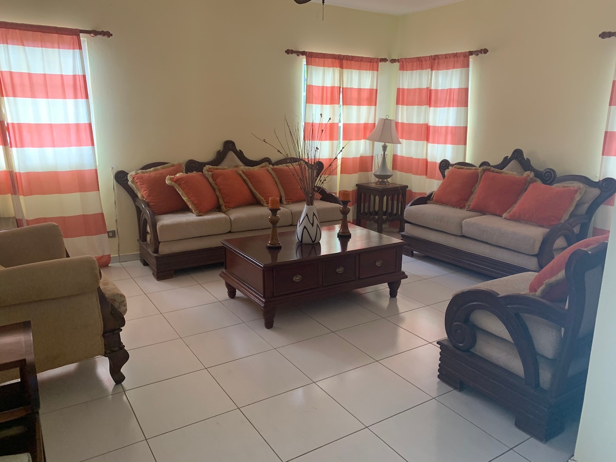 A spacious living room features two comfortable sofas with red and beige cushions, complemented by a wooden coffee table at the center. Bright striped curtains frame the windows, allowing natural light to fill the room. A lamp and decorative elements add to the welcoming ambiance.