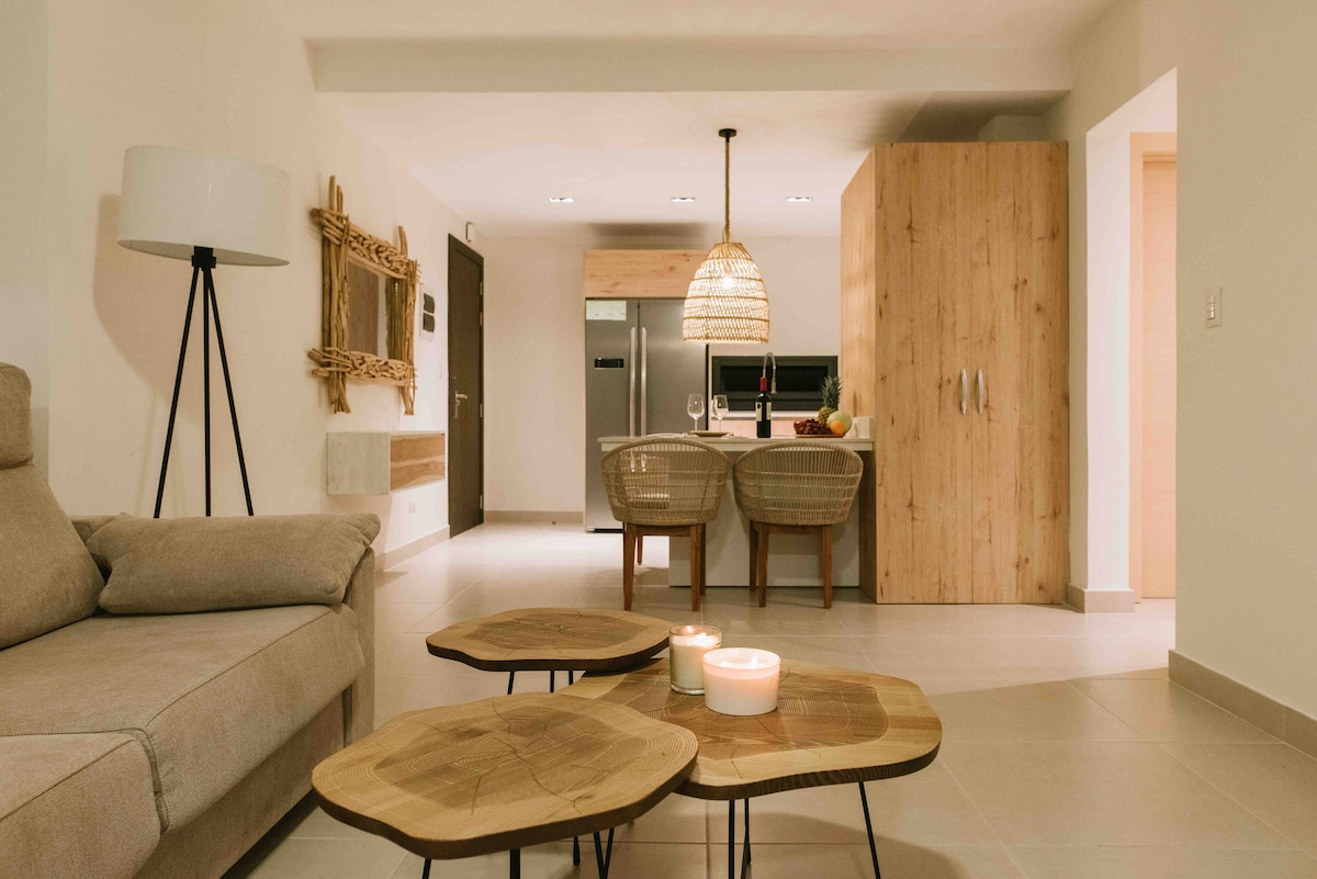 A cozy living area features a soft sofa adjacent to wooden side tables. The open kitchen showcases modern appliances and a dining space with wicker chairs. Warm lighting highlights the natural wood accents throughout the room, creating a welcoming ambiance.