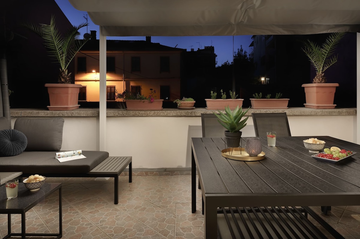 A spacious terrace is presented, furnished with a large dining table and lounge area. Potted plants line the perimeter, enhancing the relaxing atmosphere. Soft lighting from nearby buildings casts a warm glow as evening approaches, creating an inviting space for gatherings.