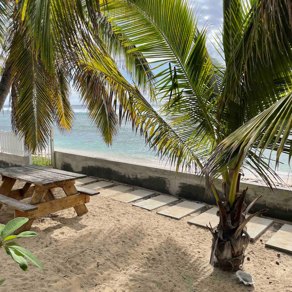 A sandy area is framed by a palm tree, leading to a clear view of the ocean. A wooden picnic table is positioned on the sand, with stone pathways visible in the background, inviting relaxation by the beach.