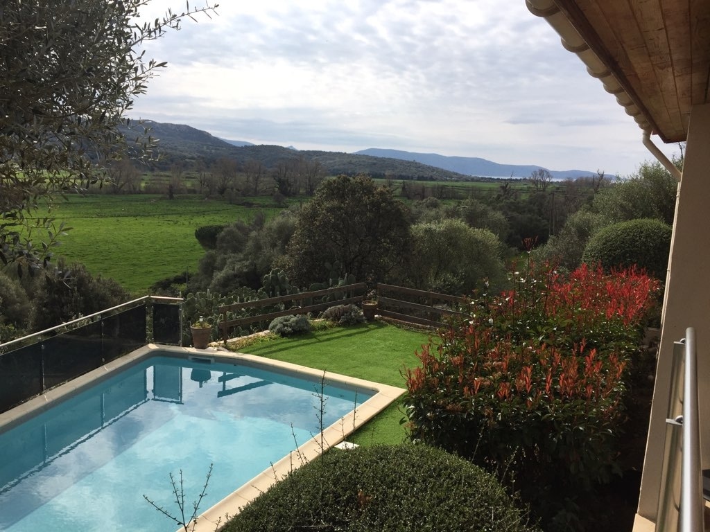 A private outdoor heated pool is framed by well-maintained gardens, showcasing vibrant green plants and colorful flowers. The expansive view reveals rolling hills and mountains under a partly cloudy sky, creating a peaceful landscape. A glass railing enhances visibility of the serene surroundings.