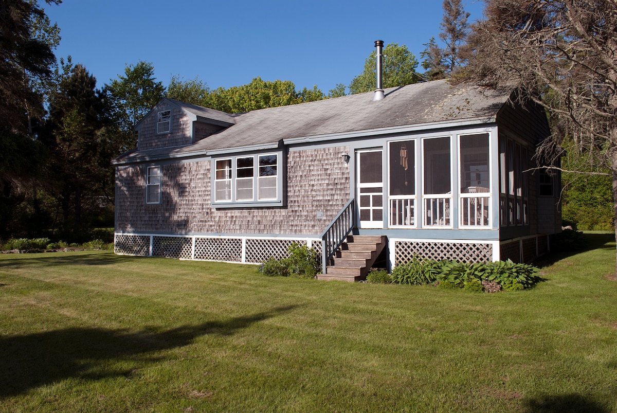 The exterior of the cottage is shown, featuring weathered shingle siding and a screened porch. Steps lead up to the entrance, with lush green grass and garden beds framed by low-growing plants. Nearby trees provide a natural backdrop under clear blue skies.