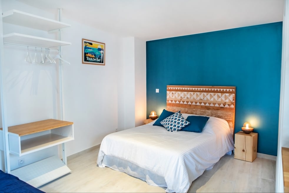 A spacious sleeping area features a double bed with a decorative headboard, complemented by side tables with lamps. A neatly organized closet with hangers is present, along with a wooden shelf. The room is illuminated by soft light, creating a calm environment.