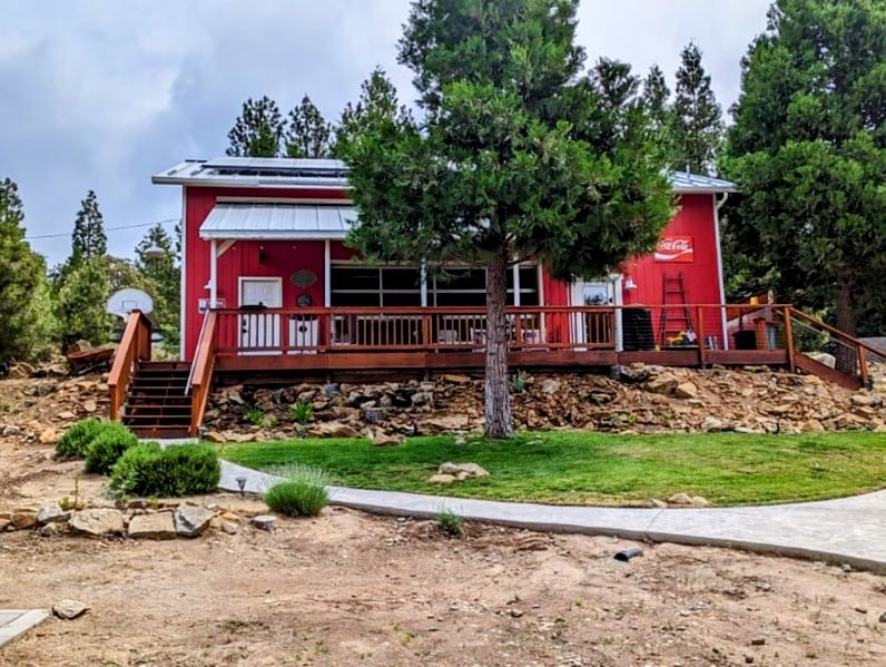 The exterior of a bright red studio is set against a backdrop of trees and rocky terrain. A wooden deck with a railing surrounds the front, featuring steps that lead down to a landscaped area with grass and shrubs. A paved path extends from the foreground.